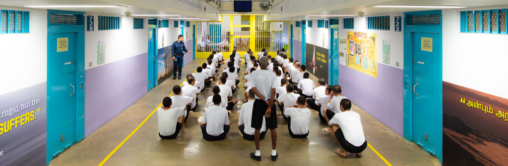 A group of inmates sitting on the ground, waiting for their prison officer to announce things.